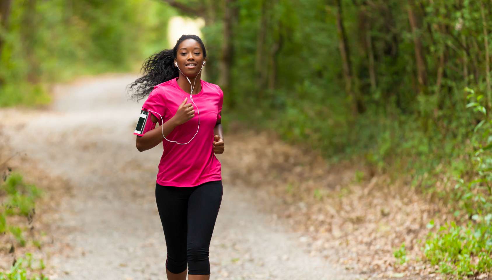 Active woman running in athletic black leggings and a pink shirt, carrying her phone in an arm band and wearing hanging earphones while enjoying a run on a dirt road surrounded by a lush forest.